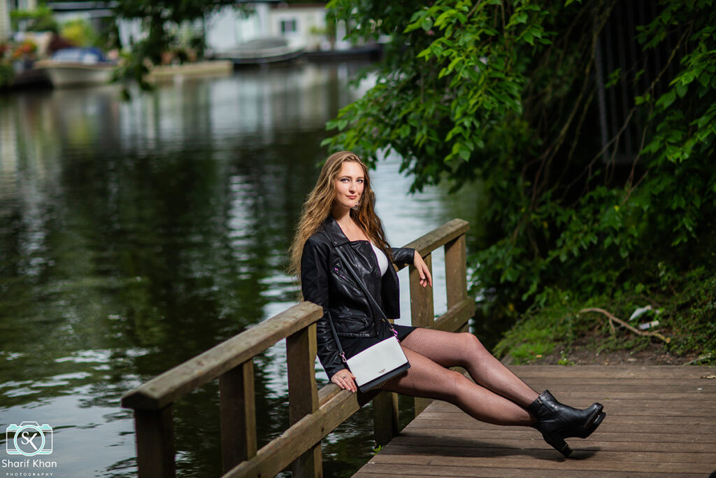 Female model Marina Krivonossova smiles at the camera during a fashion photoshoot by a canal in a city in the Netherlands. Marina leans back on the railing on which she sits.