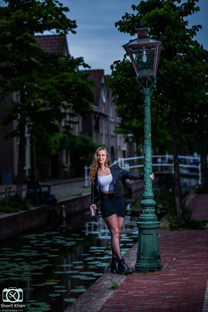 Female model Marina Krivonossova poses next to a lamp post by a canal in the Netherlands during a fashion photoshoot. Marina smiles and looks into the distance.