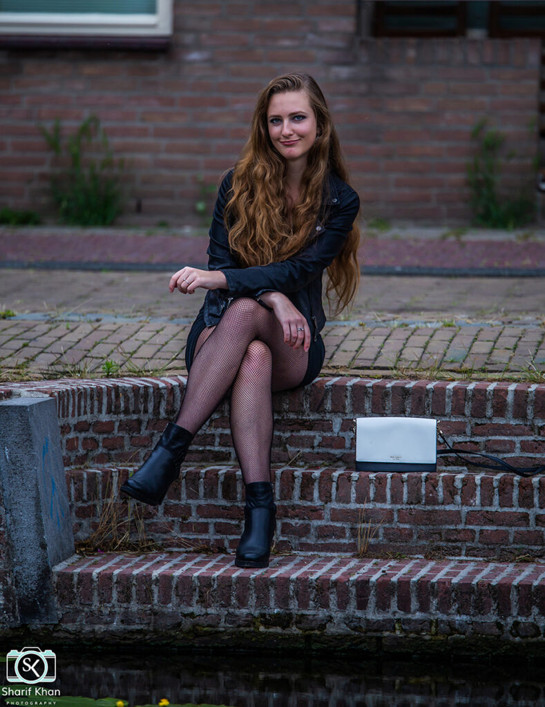 Female model Marina Krivonossova smiles at the camera during a fashion photoshoot near a canal in a city in the Netherlands. Marina crosses her legs and her arms, nonchalantly relaxing during the photoshoot.