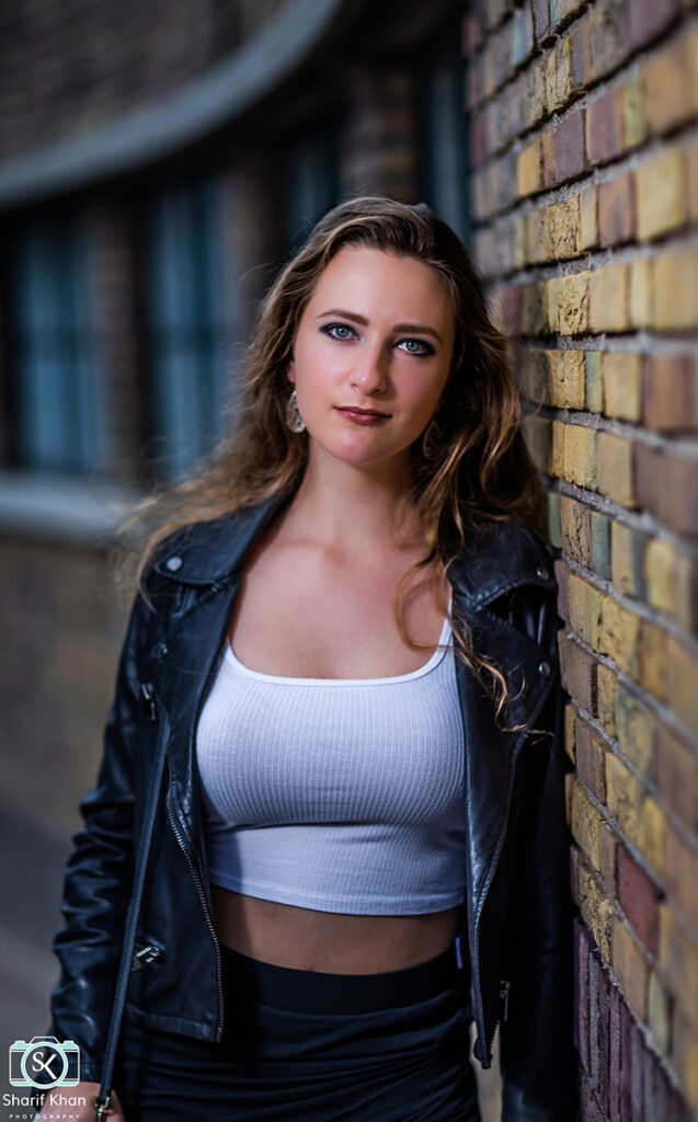 Female model Marina Krivonossova leans against a brick wall during a fashion photoshoot in a city in the Netherlands. Marina smiles slightly at the camera directly in front of her.