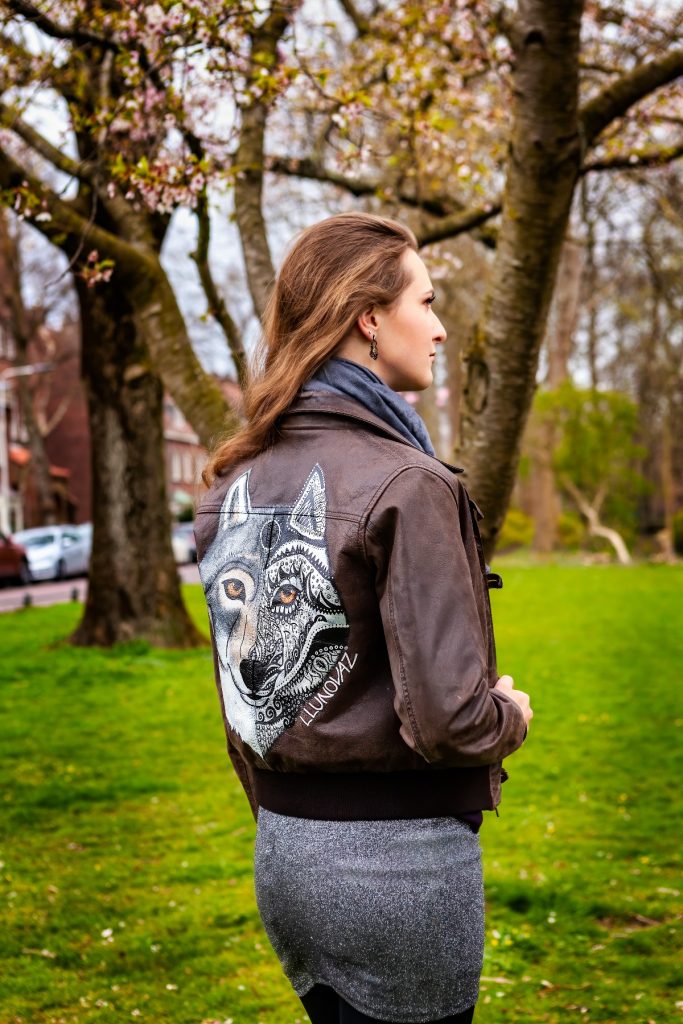 Female model Marina Krivonossova turns her back to the camera as she models a hand-painted leather jacket featuring an image of a wolf. There are beautiful trees in the background.