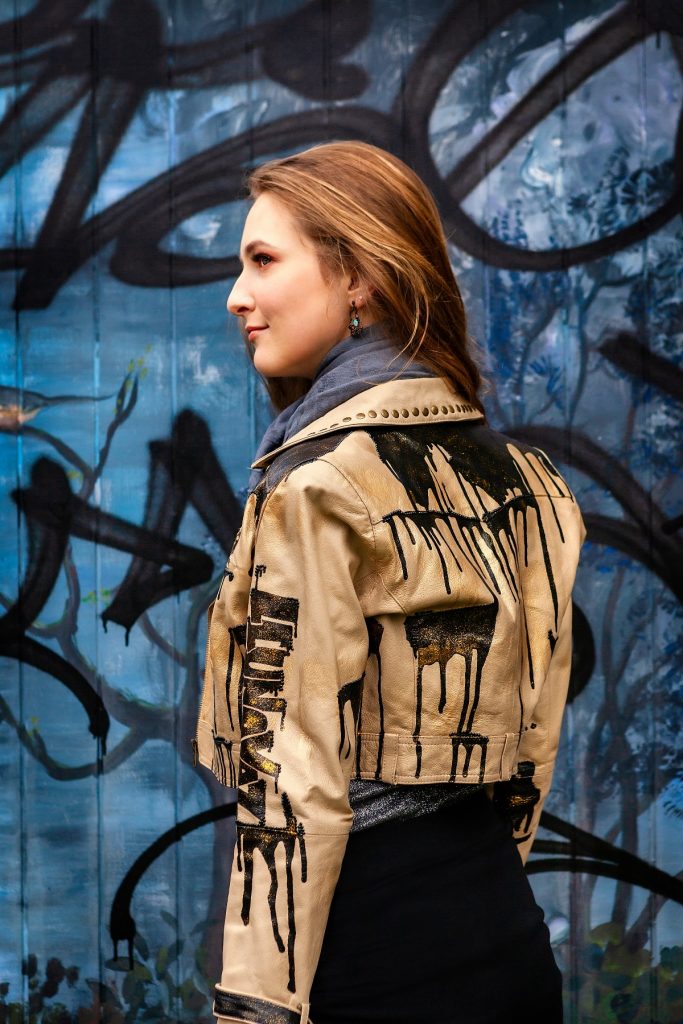 Female model Marina Krivonossova shows her back to the camera as she models a fashionable leather jacket. Marina poses in front of a graffiti wall.