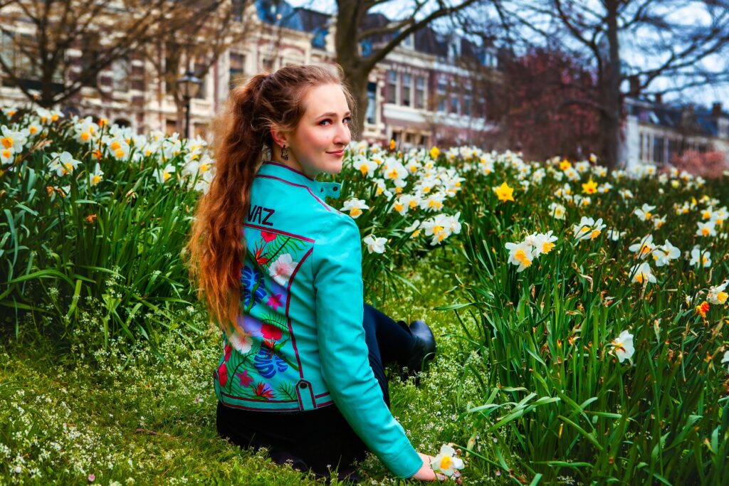 Female model Marina Krivonossova sits on the grass, surrounded by flowers, during a fashion-themed photoshoot in the Netherlands. Marina models a hand-painted leather jacket with a beautiful floral design. Marina looks over her shoulder and smiles at the camera.