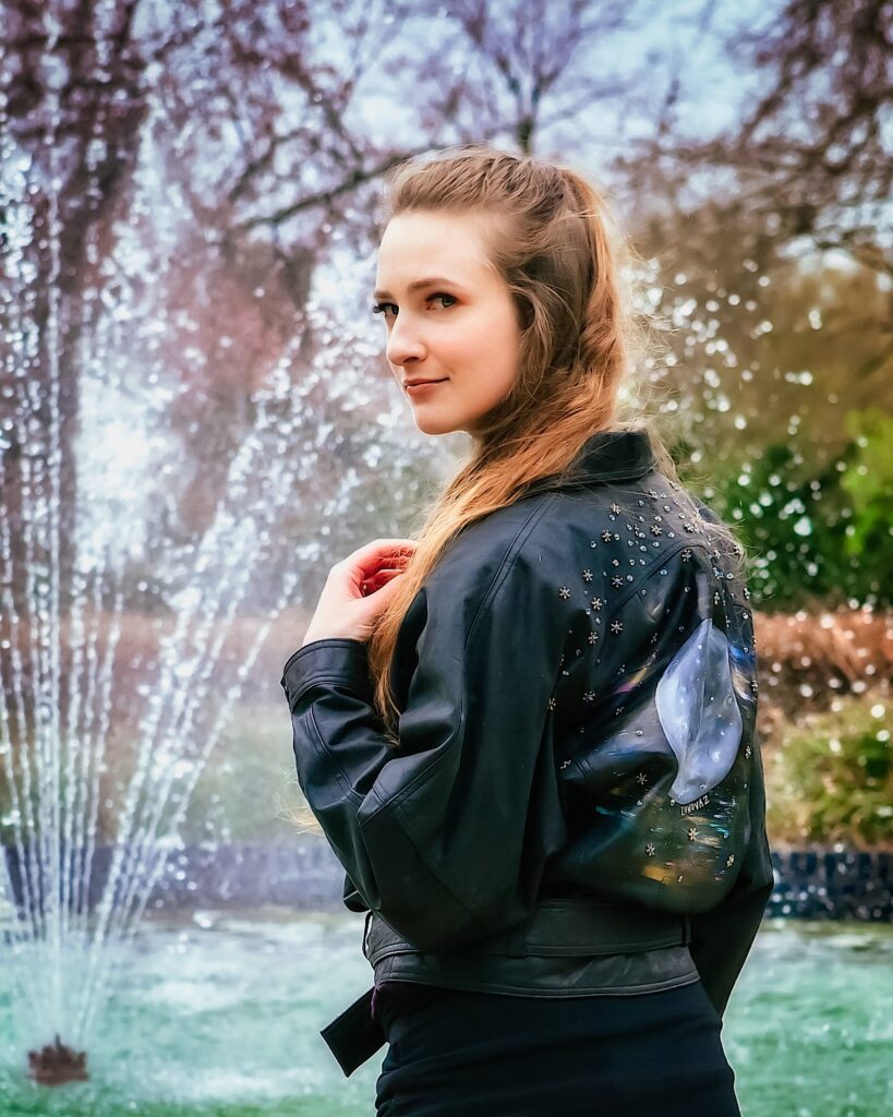 Female model Marina Krivonossova turns around and smiles slightly at the camera, all while showing off her beautiful, hand-painted leather jacket. There is a fountain in the background.
