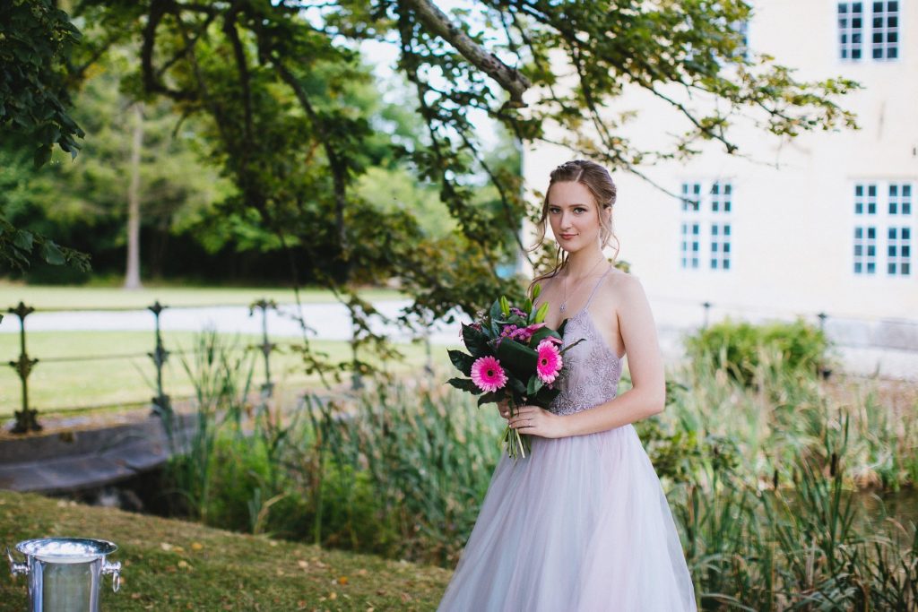 Female model photoshoot image. She looks directly at the camera during a wedding photoshoot. She holds a beautiful bouquet of flowers. There is a castle and nature in the background.
