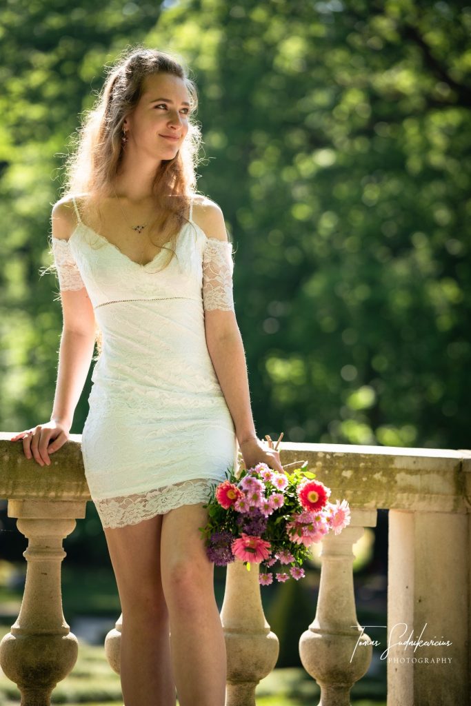 Female model photoshoot image. She holds a bouquet and looks into the distance as she smiles during a wedding photoshoot. Marina wears a traditionally white, short wedding dress for this photoshoot.