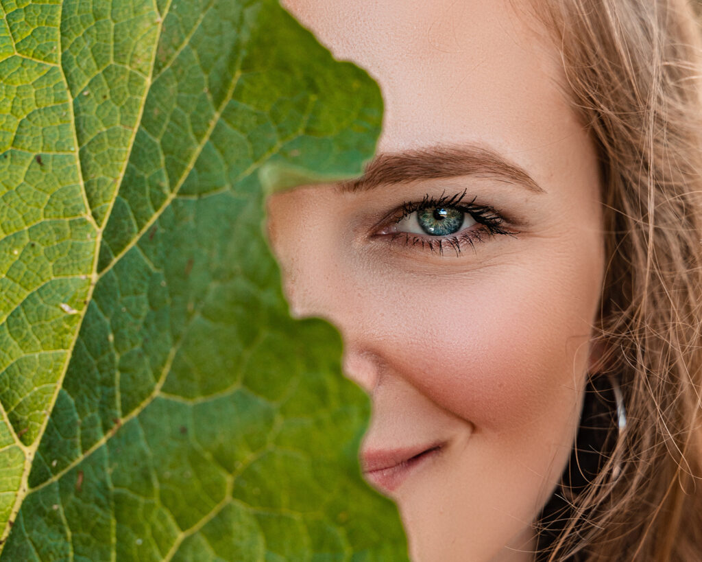 Female model Marina Krivonossova covers half of her face with a leaf and smiles at the camera during a nature photoshoot.