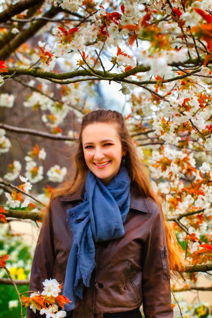 Female model Marina Krivonossova smiles at the camera during a fashion photoshoot. There is a tree with beautiful flowers in the background.