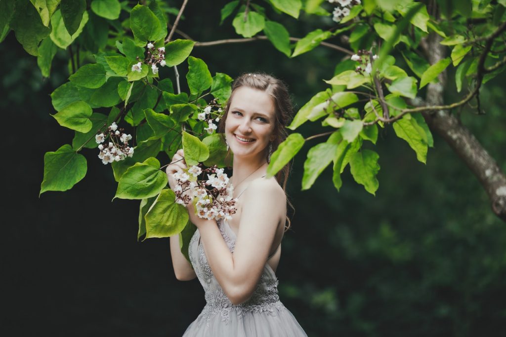Female model photoshoot image. She smiles and looks into the distance during a wedding photoshoot in Denmark. Marina holds a tree branch filled with beautiful flowers. The photoshoot takes place in a forest near a castle.